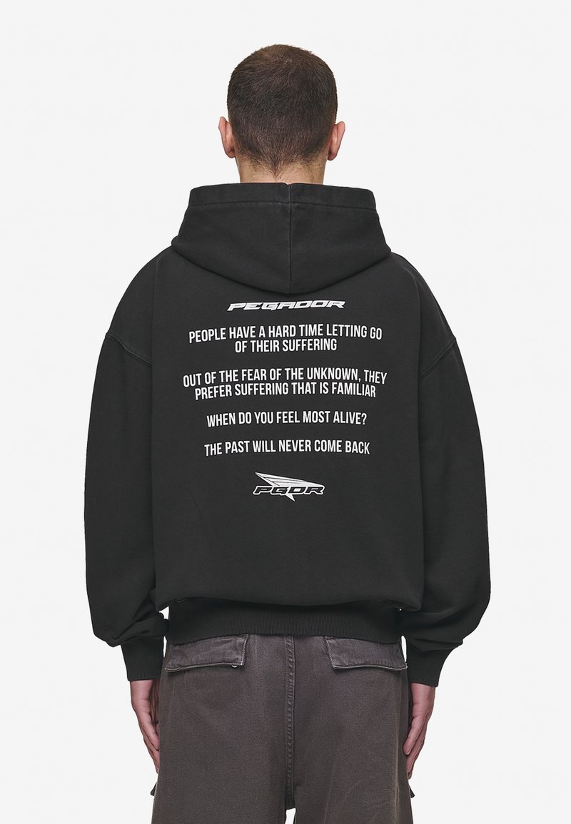 Homme portant un sweat à capuche noir avec un texte blanc au dos : phrases motivantes sur la souffrance, la peur et la vie, en police minimaliste.