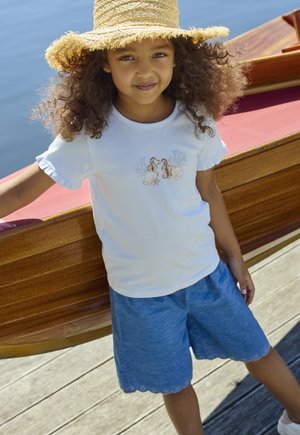 Maglietta bianca di cotone con ricamo di cavalluccio marino e maniche arruffate, abbinata a pantaloncini blu con bordi smerlati.