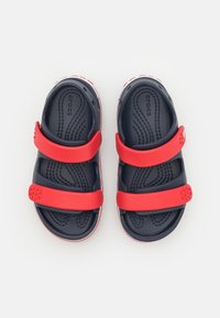 Niebieskie i czerwone sandały z dwiema regulowanymi paskami i teksturowaną wkładką. Posiadają charakterystyczne otwory wentylacyjne oraz białe akcenty na podeszwie.