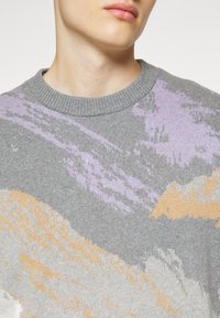 Gros plan d'une personne portant un pull gris avec des motifs abstraits lavande, beige et blanc et un col rond côtelé.