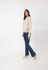 Beige gebreide cardigan met knopen, gecombineerd met donkerblauwe flared jeans en witte sandalen met bandjes met een gouden accent. Model staat zijwaarts.