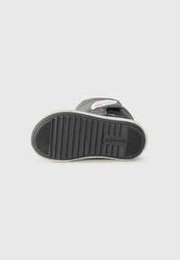 Semelle de chaussure noire avec surface texturée et rainures. Équipée d'une semelle intermédiaire blanche et d'un logo de marque coloré. Une bande Velcro visible sur le dessus.