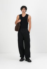 Black sleeveless tank top, loose black trousers, and brown suede shoulder bag; model stands against a white background. Black footwear with thick soles.