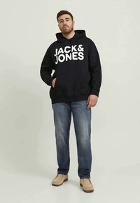 Homem a usar um moletom preto da "JACK & JONES", jeans azuis e sapatos beges, de pé com uma mão no bolso contra um fundo liso.