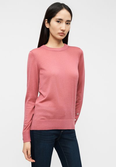 Πουλόβερ με μακριά μανίκια από απαλό, ροζ πλεκτό ύφασμα. Ριμπ λαιμόκοψη σε στρογγυλή γραμμή και πλαϊνές σχισμές, με κουμπιά διακοσμητικά στις μανσέτες. Φορεμένο με σκούρο τζιν.