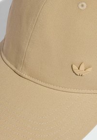 Beżowa czapka baseballowa wykonana z teksturowanej tkaniny, z małym wytłoczonym logo przedstawiającym trzy liście. Widoczne szwy wokół daszka.