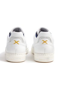 Weiße Ledersneaker mit glatter Textur, ausgestattet mit einem gelben "X"-Akzent an der Ferse und einer weißen Gummisohle. Minimalistisches Design.