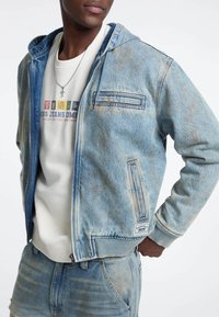 Giacca in denim con cappuccio, colore azzurro chiaro con dettagli invecchiati e tasche con zip. Indossata sopra un felpa color crema.