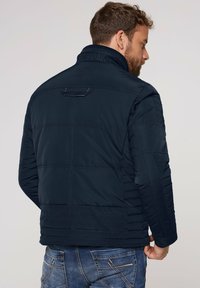 Marine pufferjakke med quiltet tekstur, høj krave og syet detaljer. Har branding på kraven og en lynlåslomme på ryggen.