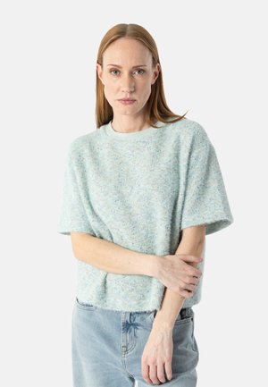 Frau mit hellbraunen Haaren, die einen hellblauen kurzärmeligen Strickpullover und hellblaue Jeans trägt, steht mit überkreuzten Armen vor einem weißen Hintergrund.