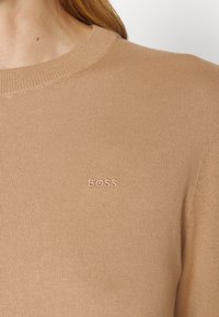 Gros plan sur une personne portant un pull en maille beige avec "BOSS" brodé sur le côté gauche de la poitrine.