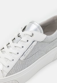 Baskets blanches et argentées avec tissu texturé, lacets blancs et un patch métallique affichant le nom de la marque sur le côté.