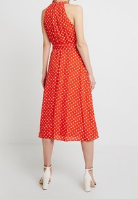 Röd klänning med polka prickar och halterneck-design, flödande tyg och midilång kjol. Bärs med vita klacksandaler.