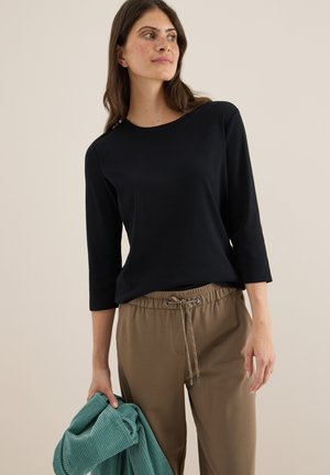 Schwarzes Langarmoberteil mit rundem Ausschnitt, kombiniert mit beigen Hosen mit elastischem Bund, hält ein türkisfarbenes Rippengewand. Schlichtes, lässiges Design.