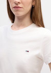 Camiseta de algodón blanca con cuello redondo, que presenta un pequeño logo bordado en rojo, blanco y azul en la parte izquierda del pecho. Textura suave.