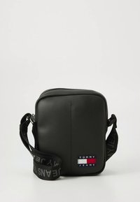 Bolso bandolera de cuero sintético negro con correa ajustable, forma rectangular, con un parche de marca en rojo, blanco y azul y diseño minimalista.
