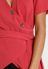 Haut croisé rouge en tissu texturé, avec manches courtes, deux boutons à l'avant et ceinture nouée à la taille.