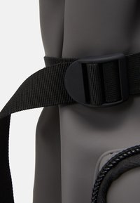 Bolsa impermeable gris con acabado texturizado, que cuenta con una correa tejido negra y un hebilla de hardware para un cierre seguro.