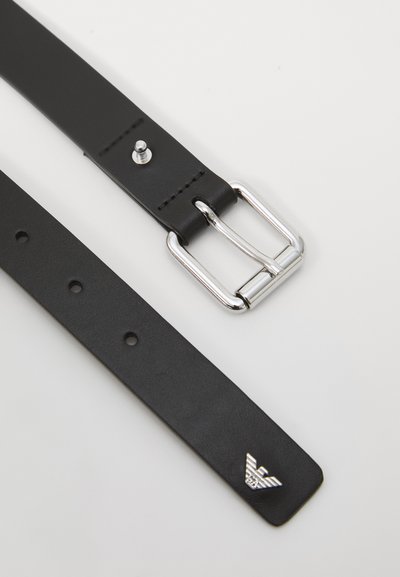 Ceinture en cuir noir avec une boucle en argent et un logo, reposant à plat sur un fond clair, mettant en valeur son design élégant et ses fermetures.