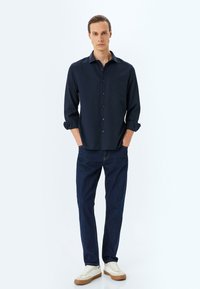 Mörkblå knapp-up skjorta, upp- och nedvikta ärmar, ihop med blå denimjeans och beige sneakers. Tyget verkar slätt med minimal textur.