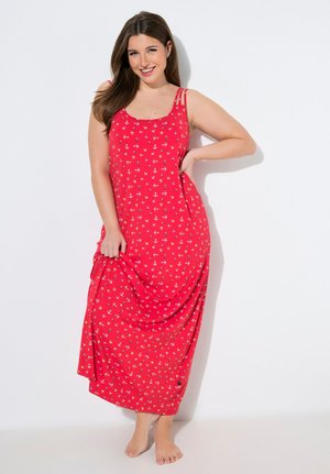 Mujer sonriente con un vestido maxi rojo sin mangas con un patrón de anclas blancas, de pie descalza contra una pared blanca.