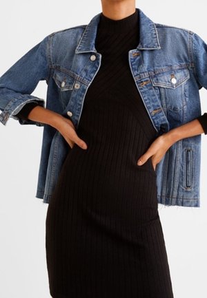 Persona con chaqueta vaquera azul sobre un vestido negro ajustado de canalé, de pie con las manos en las caderas frente a un fondo liso.