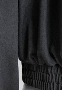 Svart tyg med en slät yta, med en rynkad kant och manschetter. Designen betonar strömlinjeformade, minimalistiska element.