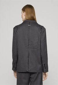 Svart blazer med tynne striper og tilpasset passform, med hakket krage og knappede mansjetter, teksturert stoff og subtil logo-brodering på baksiden.