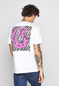 White t-shirt featuring a pink smiley face graphic on a zebra-striped black and white background, short sleeves, and a crew neck design.