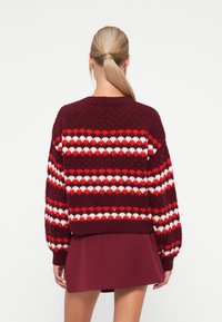 Maroon knitted sweater with red and white heart patterns, wide sleeves, and a ribbed hem. Model is wearing a burgundy skirt.