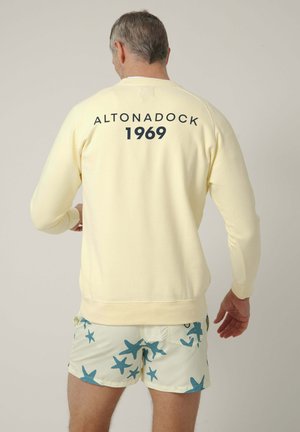 Hombre con sudadera amarillo pálido con "ALTONADOCK 1969" en la espalda y pantalones cortos claros con patrón de estrellas de mar azules, de pie contra un fondo liso.
