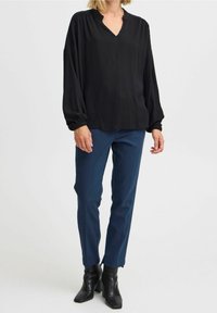 Kvinna i en lös svart blus med samlade ärmar, figursydda marinblå byxor och svarta klackskaft på en vit bakgrund.