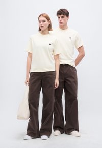 Μπεζ βαμβακερά μπλουζάκια με μικρό μαύρο λογότυπο, συνδυασμένα με σκούρα καφέ παντελόνια φαρδιάς γραμμής, συνοδευόμενα από ένα ανοιχτό μπεζ τσαντάκι. Λευκά παπούτσια.