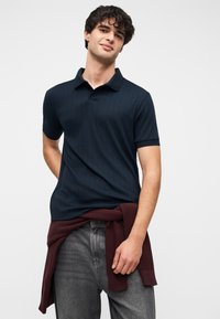 Polo bleu marine côtelé avec col, manches courtes, et un pull en maille bordeaux noué autour de la taille, assorti à un jean foncé.