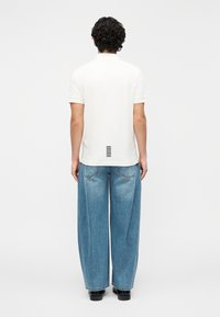 T-shirt branca de manga curta com detalhe de risca preta nas costas, combinada com jeans de perna larga azul clara. Fundo liso.