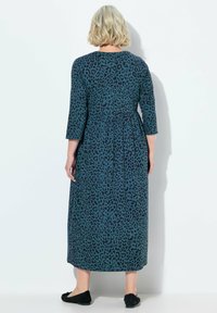 Robe longue turquoise avec un imprimé léopard noir, manches trois quarts, taille froncée et longueur jusqu'aux chevilles. Portée avec des chaussures plates noires.