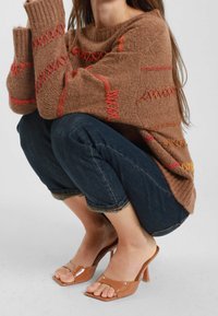 Maglione lavorato a maglia marrone con vestibilità morbida, caratterizzato da cuciture orizzontali rosse e arancioni. Abbinato a jeans blu scuro con risvolto e mule color carne.
