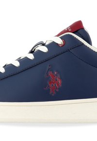 Baskets bleu marine avec lacets blancs, semelle blanche, languette rouge au talon et logo brodé rouge d’un joueur de polo sur le côté.