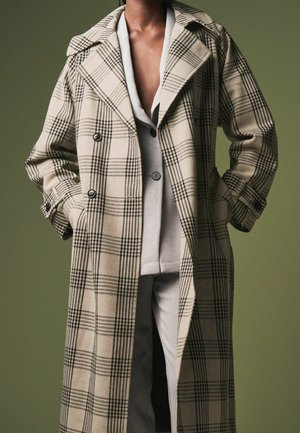 Beige ternet trenchcoat med dobbeltradet design, store udskårne revers og sidelommer, båret over et lysegrå outfit.