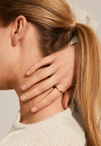 Anillo dorado ondulado en una mano, descansando sobre un cuello. Cabello castaño claro y suéter blanco texturizado visibles, con un fondo suave y neutral.