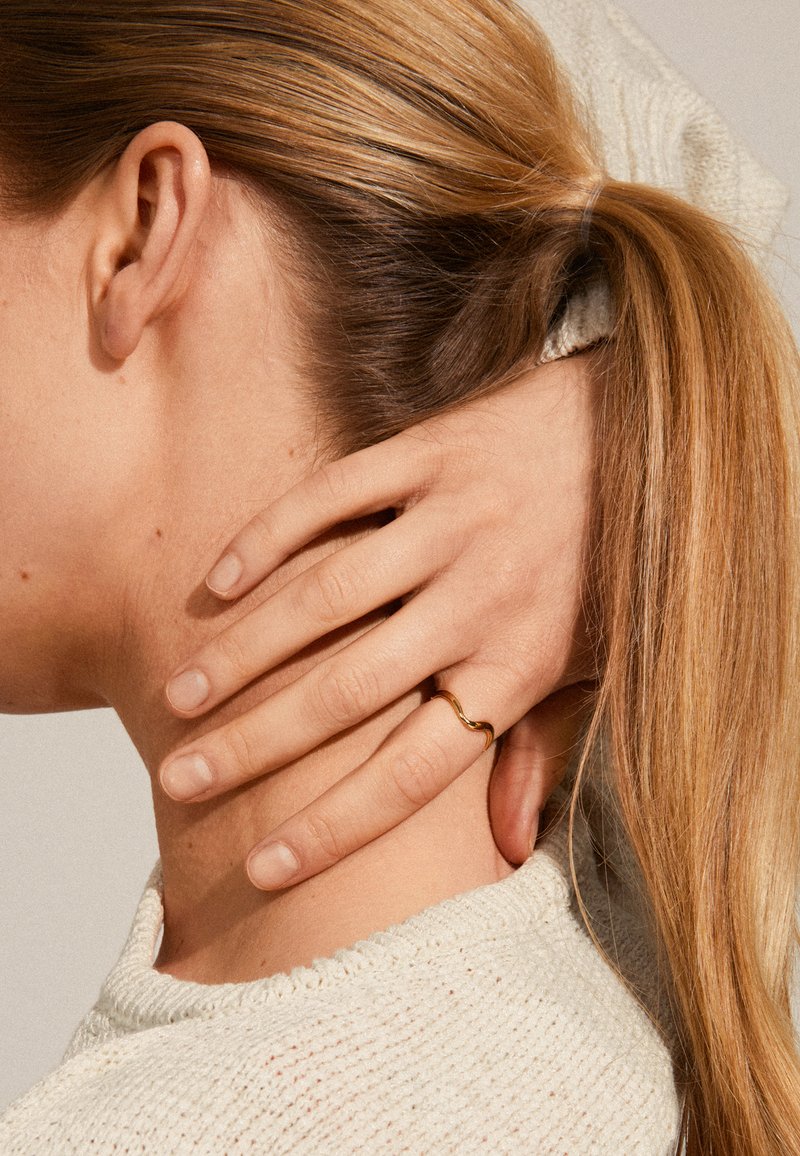 Anillo dorado ondulado en una mano, descansando sobre un cuello. Cabello castaño claro y suéter blanco texturizado visibles, con un fondo suave y neutral.
