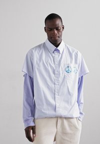 Camicia a maniche corte a righe in azzurro chiaro e bianco, con una tasca decorata con ricamo del simbolo della pace blu. Indossata sopra una camicia a maniche lunghe lavanda.