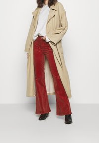 Un trench beige sur un sweat gris, associé à un pantalon en velours côtelé rouge à pattes d'éléphant et des bottes à talons noires, sur fond blanc.