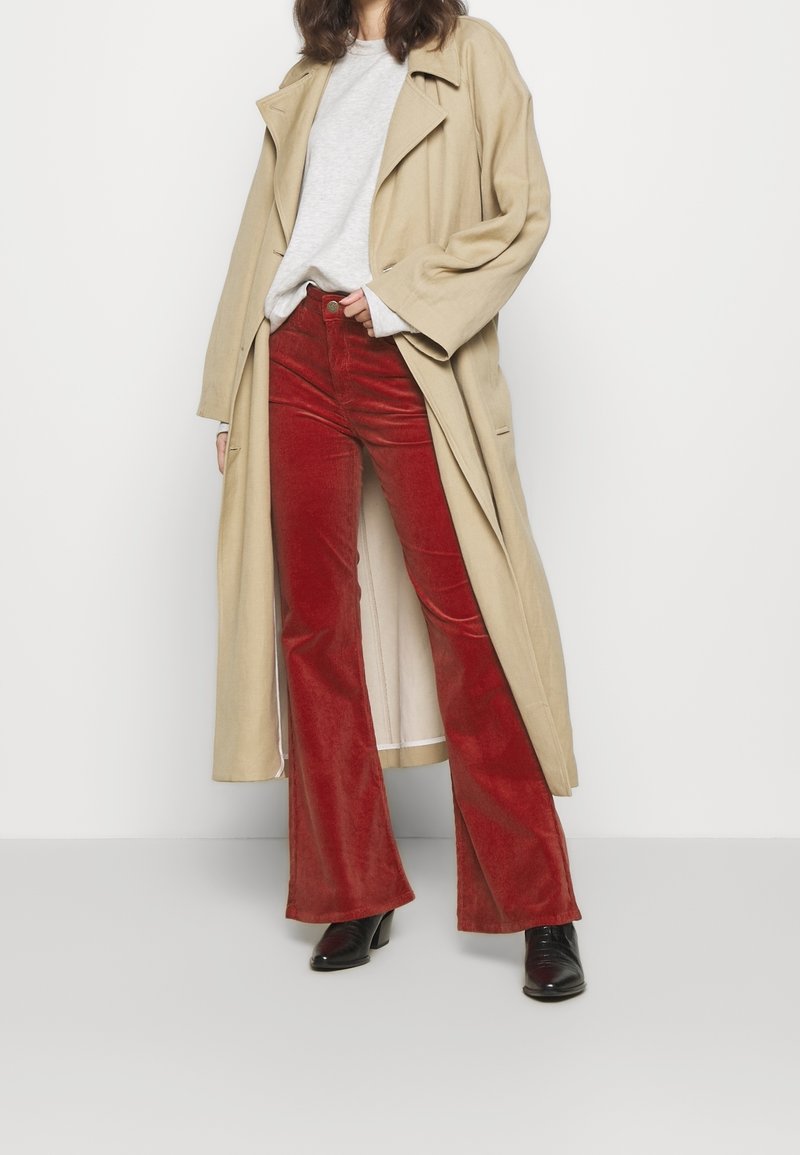 Un trench beige sur un sweat gris, associé à un pantalon en velours côtelé rouge à pattes d'éléphant et des bottes à talons noires, sur fond blanc.