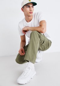 Khaki cargo nadrág, fehér póló és fehér sportcipő. A sapka bézs színű, zöld karimával és sárga logóval az elején.