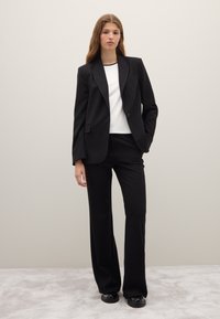 Blazer nero su misura con un solo bottone, abbinato a pantaloni coordinati e una maglietta bianca. Tessuto liscio, vestibilità strutturata, design minimalista.