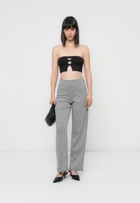 Haut noir sans bretelles avec trois découpe à l'avant, associé à un pantalon ample à carreaux gris et noir. Le mannequin tient un sac à main noir.