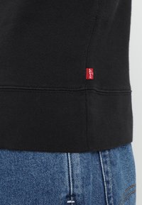 Sudadera negra con dobladillo acanalado y una pequeña etiqueta roja de Levi's en un lado, combinada con jeans azules que muestran una textura de denim desgastado.
