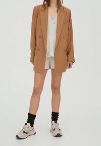 Blazer marron croisé porté sur un haut texturé clair et un short, associé à des baskets beiges et des chaussettes foncées côtelées.