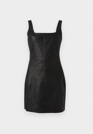 Nainen, jolla on päällään hihaton musta nahkainen minimekko ja mustat keskivartta peittävät saappaat, seisoo yksiväristä valkoista taustaa vasten.